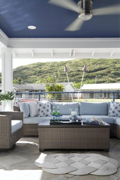 Outdoor patio on the lanai of a new home, featuring wicker furniture with light blue cushions, a black coffee table with drinks, decorative pillows, and a ceiling fan, set against scenic Oahu hills and houses.