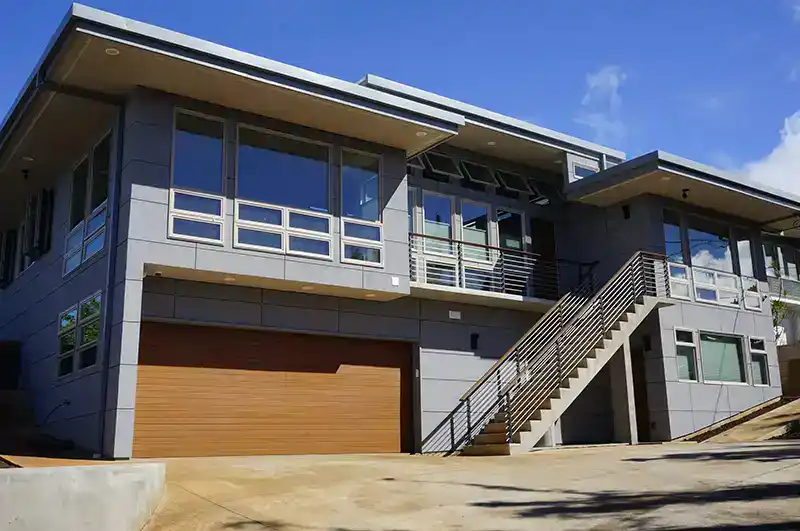 Modern two-story home with large windows, a flat roof, a wooden double garage door, and an exterior staircase leading to the upper floor. The sky is clear and blue.